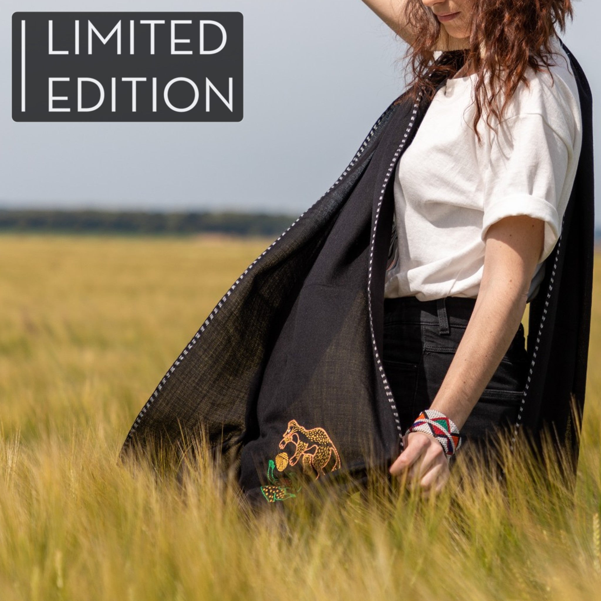 Femme dans un champ de blé portant une étole en pashmina noire brodée réalisée en collaboration avec Laïte Works. Woman in a wheat field wearing a black embroidered pashmina stole, in collaboration with Laïte Works.